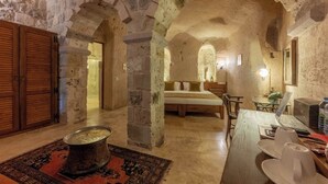 A stone-walled room with a bed, a table with a laptop, and a dining area with a table and chairs. - Peristyle Cave Cappadocia - Special Class (Nevsehir)