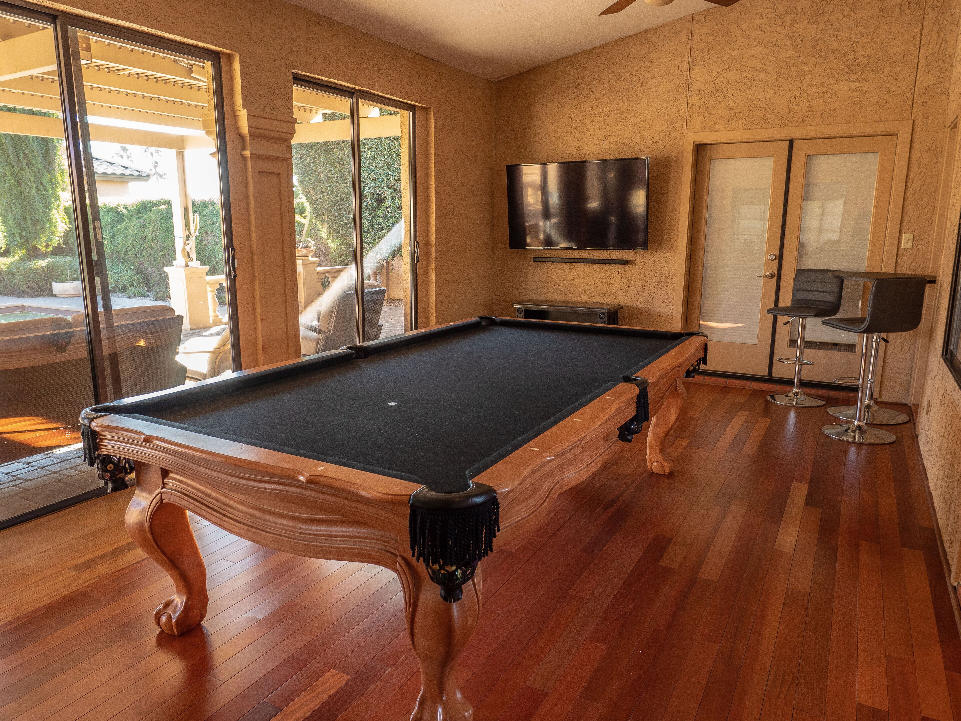 A pool table with a wooden frame and a black felt surface, a television mounted on the wall, and a seating area with two bar stools.