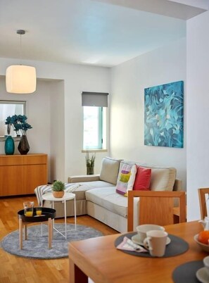 Un salon moderne avec un canapé, une petite table avec des tasses, une chaise en bois, un tapis gris et un tableau accroché au mur.