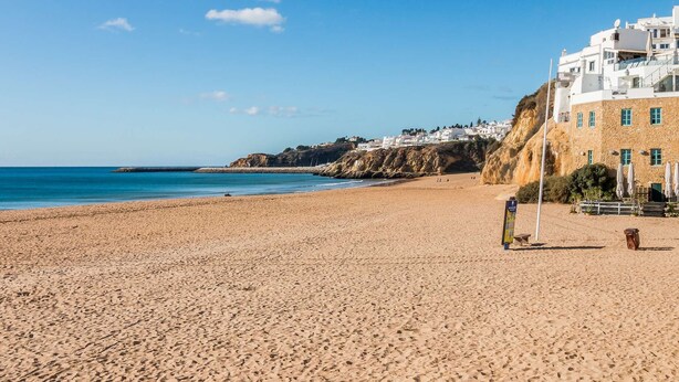 A beach with a sandy shore, a clear blue sky, and buildings in the background. - ? Elegant 1-Bedroom Apartment | Pool | Walk to Old Town & Fisherman’s Beach (Albufeira)