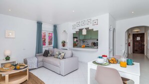 A modern living room with a grey sofa, a dining table with chairs, and a kitchen area with green cabinets. - ? Elegant 1-Bedroom Apartment | Pool | Walk to Old Town & Fisherman’s Beach (Albufeira)