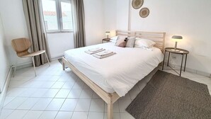A bedroom with a wooden bed, two bedside tables with lamps, a chair, a window with curtains, and a wall-mounted air conditioner. - ? Elegant 1-Bedroom Apartment | Pool | Walk to Old Town & Fisherman’s Beach (Albufeira)