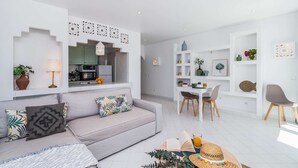A modern living room with a grey sofa, a dining area with a white table and chairs, and a kitchen with green cabinets. - ? Elegant 1-Bedroom Apartment | Pool | Walk to Old Town & Fisherman’s Beach (Albufeira)