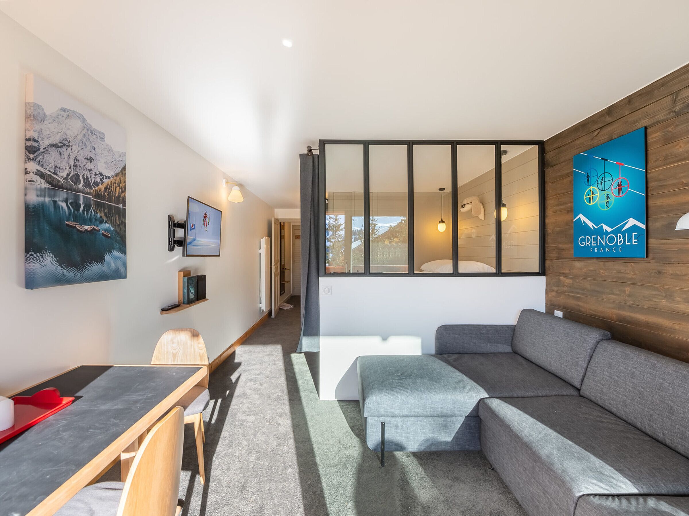A modern living room with a grey sofa, a dining table with chairs, a TV mounted on the wall, and a large window with a view of the outdoors.