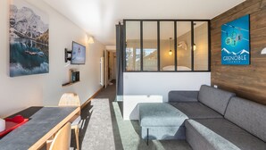 Una sala moderna con un sofá gris, una mesa de comedor con sillas, un televisor montado en la pared y un ventanal con vistas al exterior.