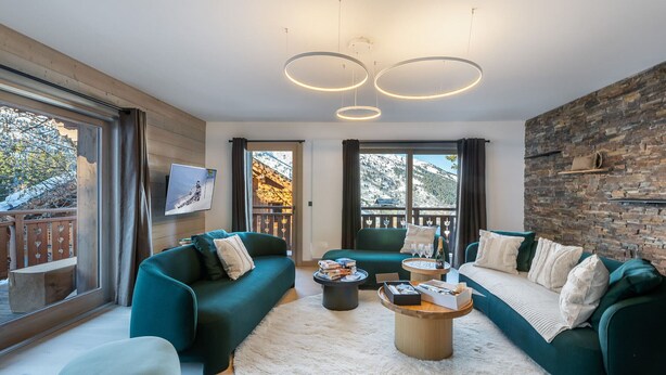 Una sala moderna con dos sofás verdes, una mesa redonda de centro y un amplio ventanal con vistas a una montaña nevada.