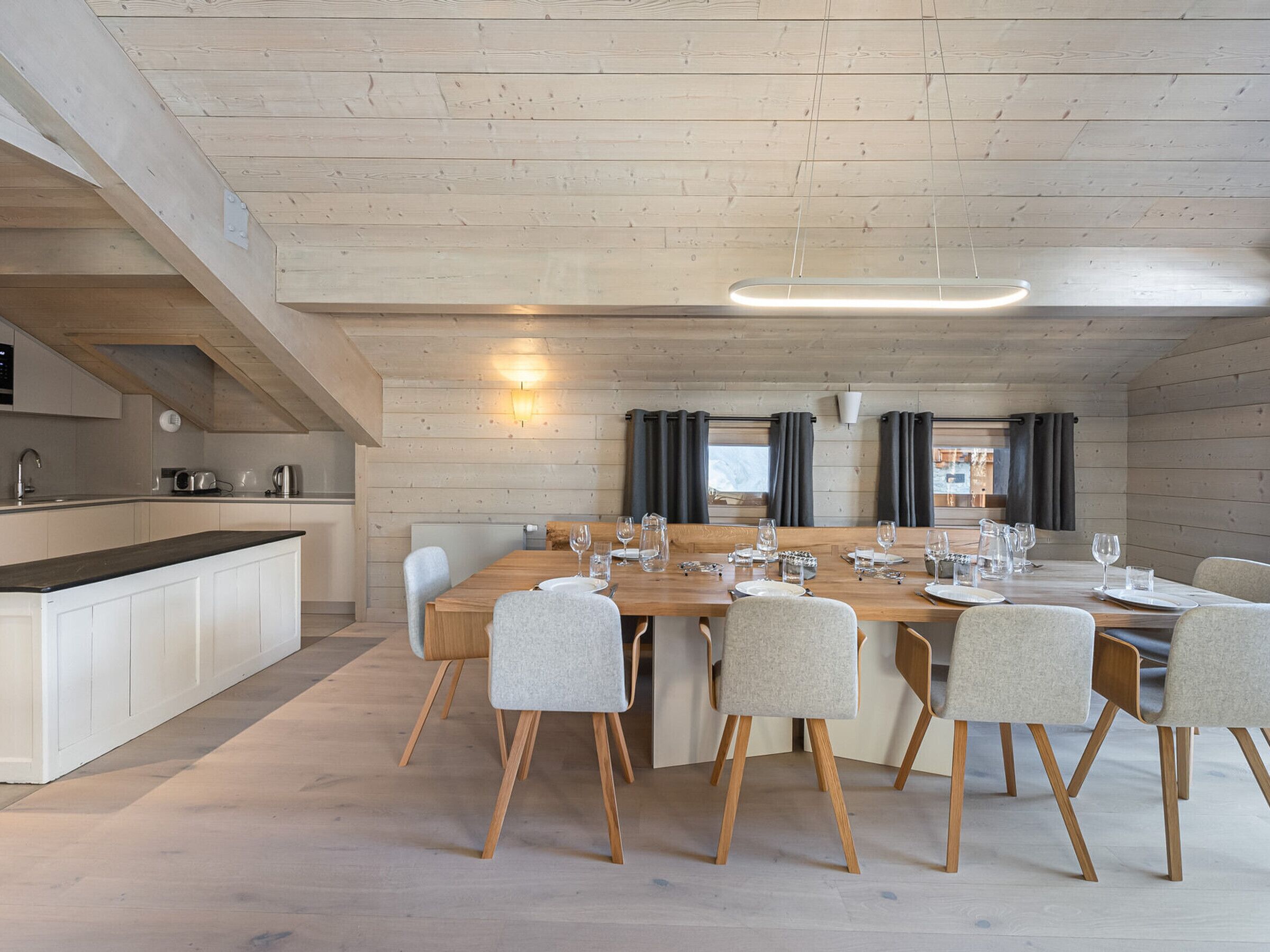 A modern kitchen with a wooden table set for dining, a built-in oven, and a large window with curtains.
