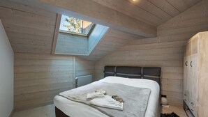 Une chambre avec un plafond en pente, un puits de lumière, un lit recouvert d’une couverture grise, un radiateur et une armoire.