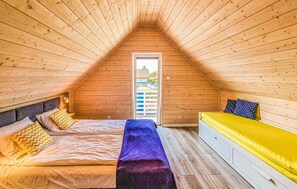 Een knus zolderkamer met schuine wanden, een bed met een paarse deken, een gele bank en een raam met uitzicht op een huis.