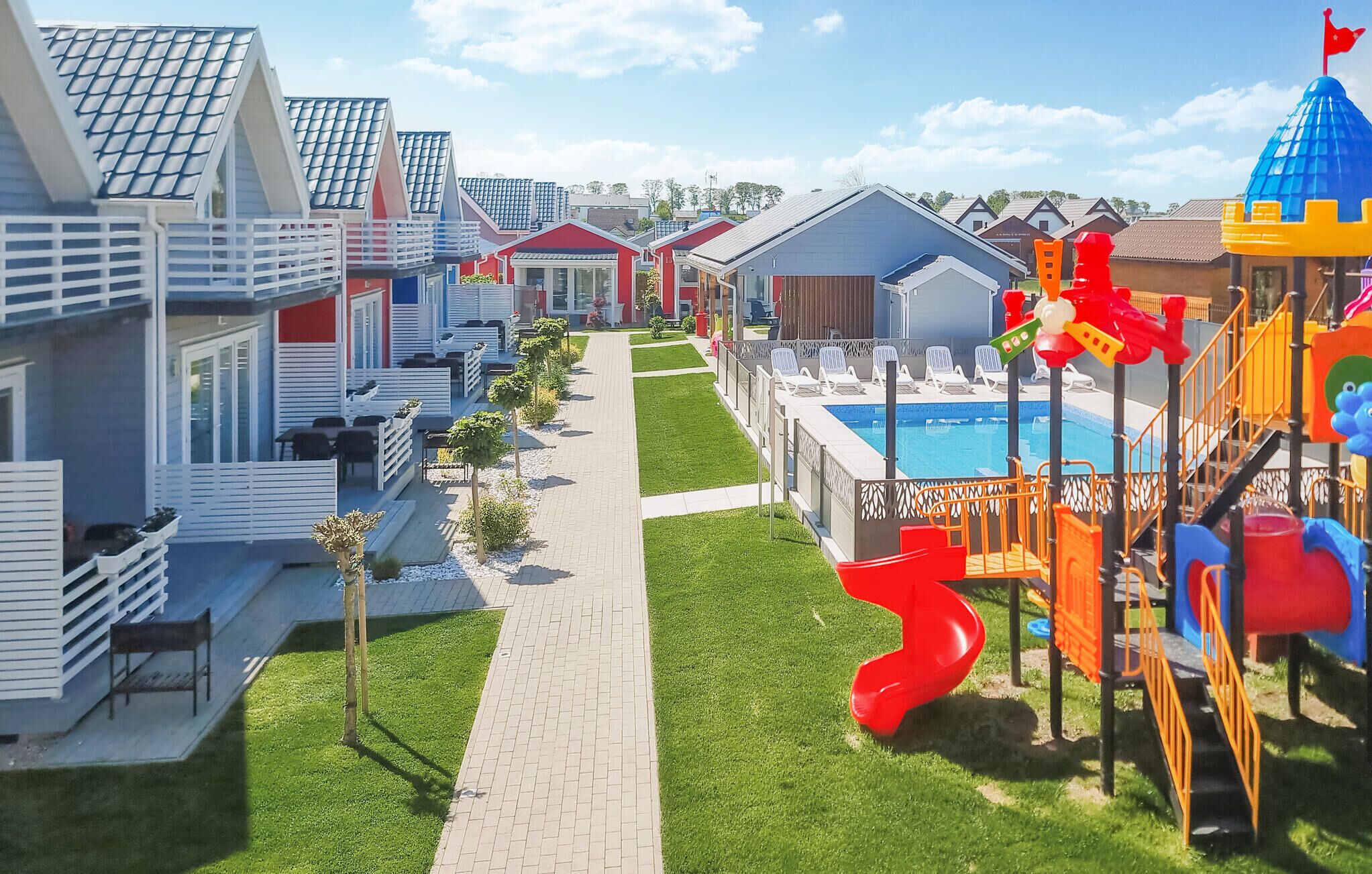 Et resort med en central sti, udendørs siddepladser, et poolområde og et legehus til børn.