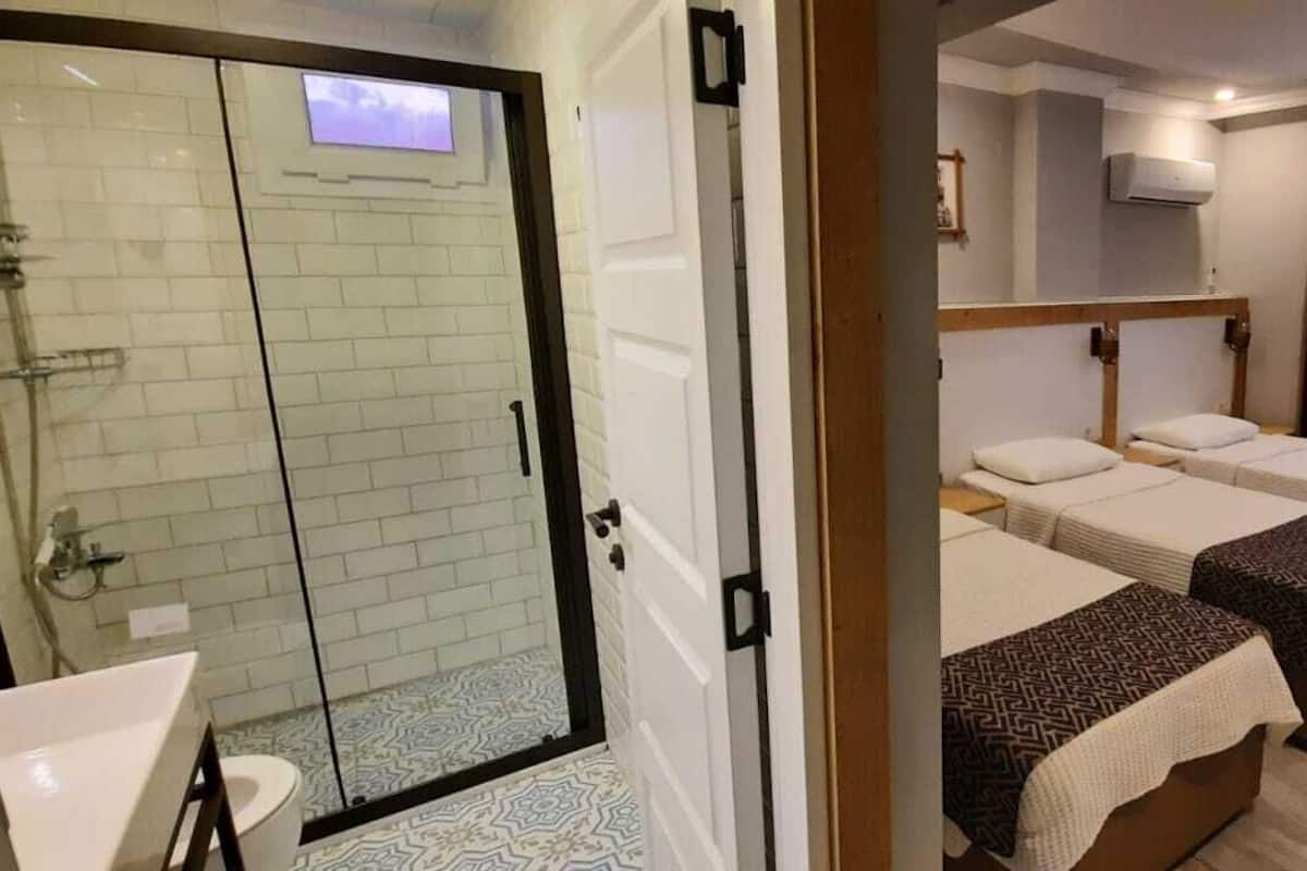 Habitación de hotel con cabina de ducha de vidrio, dos camas con cubrecamas estampados, una mesita de noche y un televisor montado en la pared.