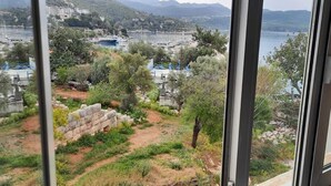 A view from a window overlooking a coastal area with boats, greenery, and a distant town. - Yalı Pansiyon (Kas)