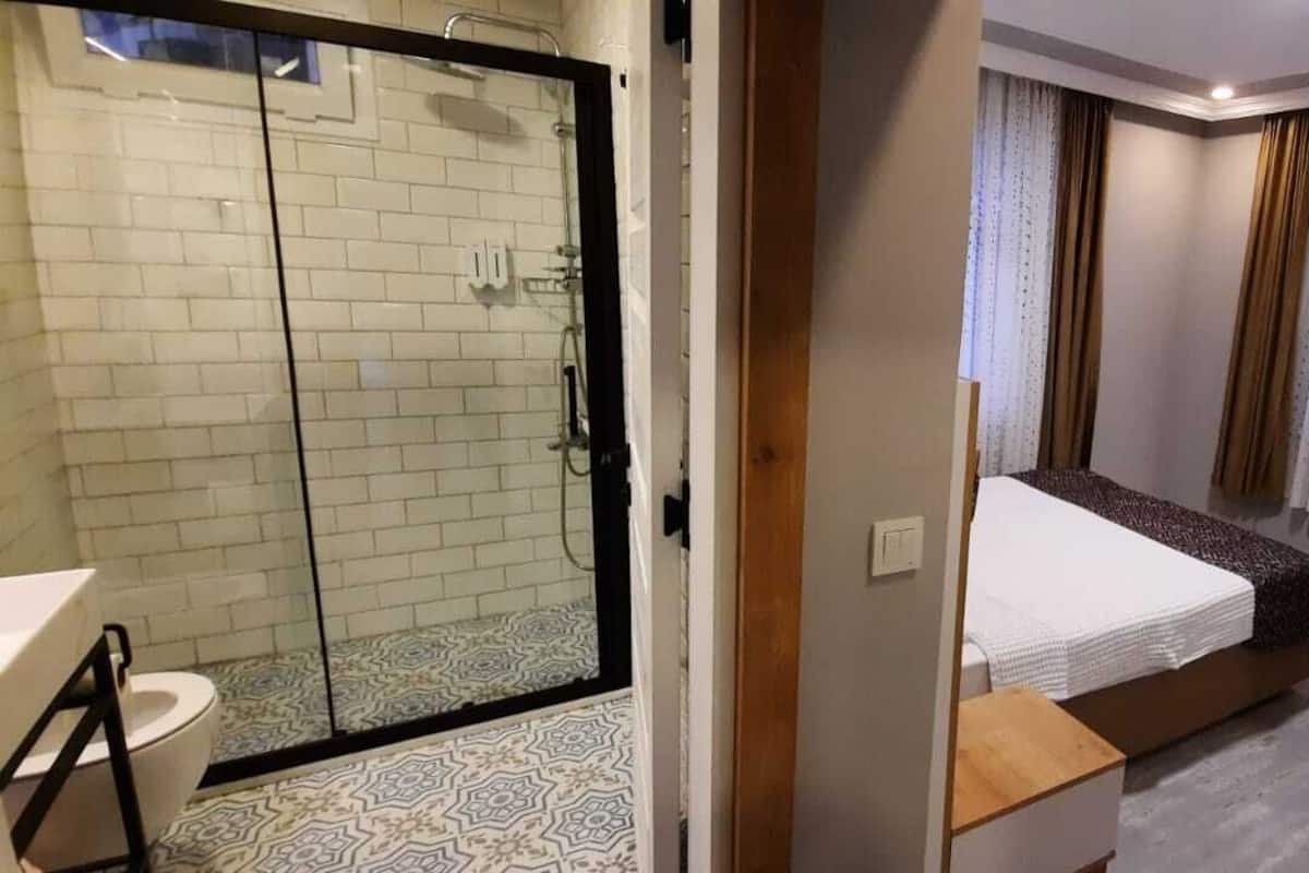 Habitación de hotel con cabina de ducha de vidrio, cama con colcha blanca y marrón, mesita de noche pequeña, una silla y televisor sobre un mueble.