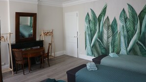 Une chambre à coucher comprenant un lit, un bureau, une chaise, un miroir et un mur orné d’une grande fresque représentant des feuilles.