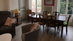 Un salon chaleureux avec un canapé aux couleurs sombres, une petite table d’appoint avec une lampe, une table à manger avec des chaises, et une grande fenêtre donnant sur des arbres et un immeuble.