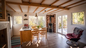 A cozy kitchen with wooden beams, a dining area with a table and chairs, a fireplace, and large windows offering views of the outdoors. - Sunny Swedish cottage surrounded by forest & tranquillity (Åsensbruk)
