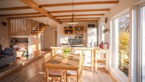 A spacious kitchen with wooden beams, a dining area with a table and chairs, and a view of autumn foliage through large windows. - Sunny Swedish cottage surrounded by forest & tranquillity (Åsensbruk)