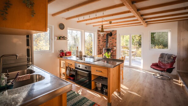 A modern kitchen with a central island, wooden beams, large windows, and a fireplace. - Sunny Swedish cottage surrounded by forest & tranquillity (Åsensbruk)