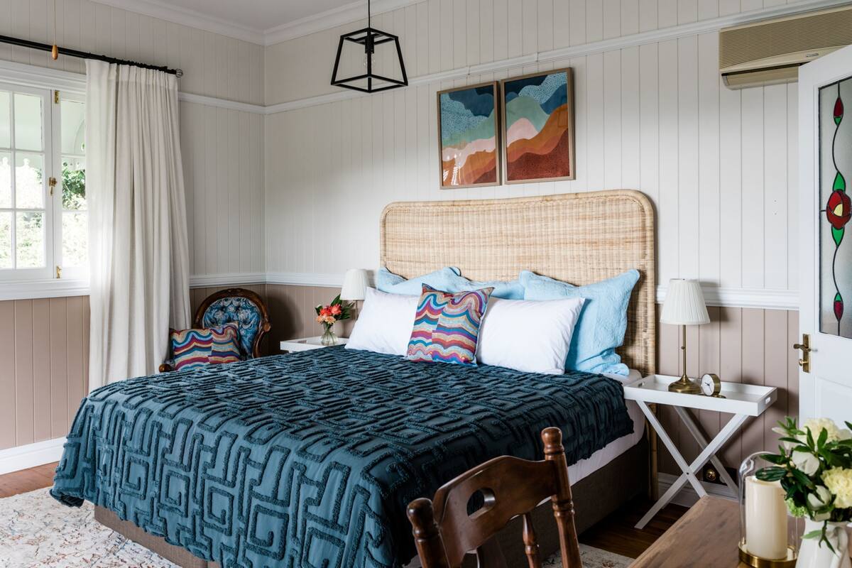 Une chambre à coucher comprenant un lit, des tables de chevet, une chaise, une fenêtre avec des rideaux et des œuvres d’art murales.