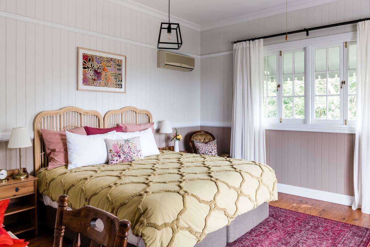 Une chambre avec un grand lit, une table de chevet avec une lampe, une chaise, une fenêtre avec des rideaux et un appareil de climatisation.