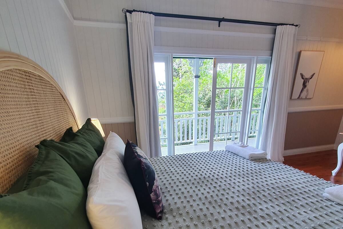 Un lit doté d’un matelas texturé, de coussins verts et blancs, et offrant une vue sur un balcon à travers une fenêtre ornée de rideaux blancs.
