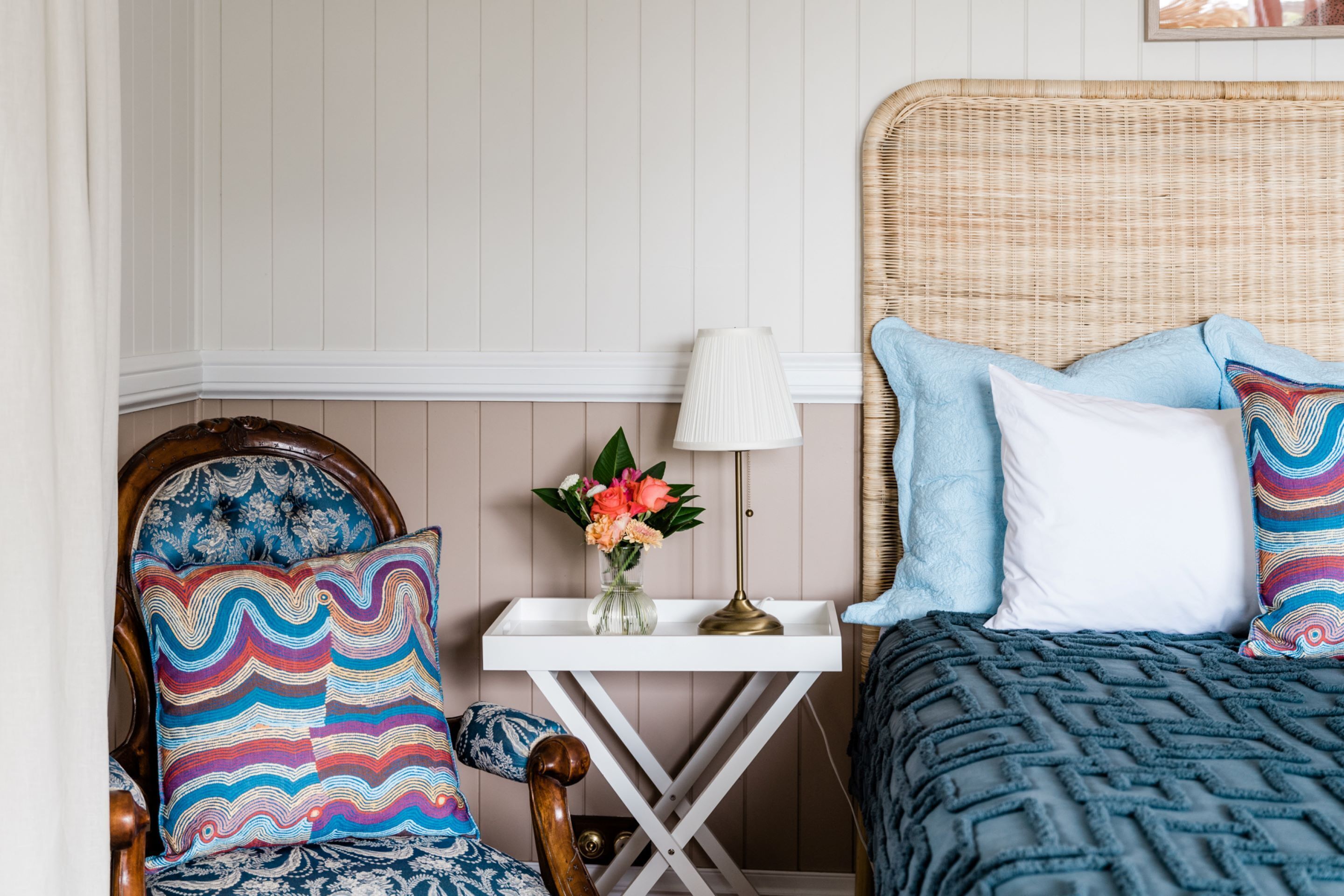 A bedroom with a wooden chair, a bedside table with a lamp, and a bed with patterned pillows.