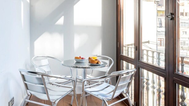Un balcon avec une table ronde et quatre chaises, un saladier rempli de fruits et un verre de jus d’orange.