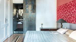 Une chambre d’hôtel moderne avec un grand lit, un mur décoré d’un motif rouge, une grande colonne et une salle de bain visible à travers une porte ouverte.