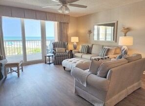 Ein geräumiges Wohnzimmer mit großem Fenster, Deckenventilator und Blick auf den Strand.