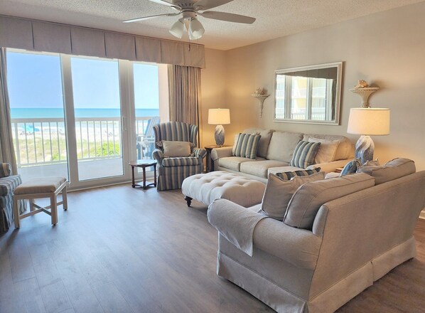 A spacious living room with a large window, a ceiling fan, and a view of the beach. - Warwick 203 Oceanfront - Great views of Ocean & Pool, Clubhouse Access (Pawleys Island)