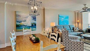 Un coin repas avec une table en bois et des chaises blanches, un tableau représentant un paysage marin et, au fond, un canapé moderne.