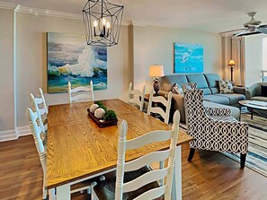 Un coin repas avec une table en bois et des chaises blanches, un tableau représentant un paysage marin et, au fond, un canapé moderne.