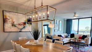 Een moderne woonkamer met een eethoek, met een houten eettafel, witte stoelen, een groot abstract schilderij en een uitzicht op de natuur door grote ramen.