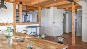 Een houten eettafel met stoelen, een vaas en een metalen mand. Achtergrond toont een keuken met een koelkast en een spoelbak.