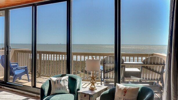 A balcony with a view of the ocean, furnished with two green armchairs, a small table with a lamp, and a chair. - Steps To The Sea | Penthouse with Ocean Views (Port Aransas)