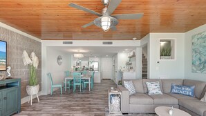 A spacious living area with a wooden ceiling, a dining area with turquoise chairs, and a gray sofa with a 'Some to the Beach' pillow. - Steps To The Sea | Penthouse with Ocean Views (Port Aransas)