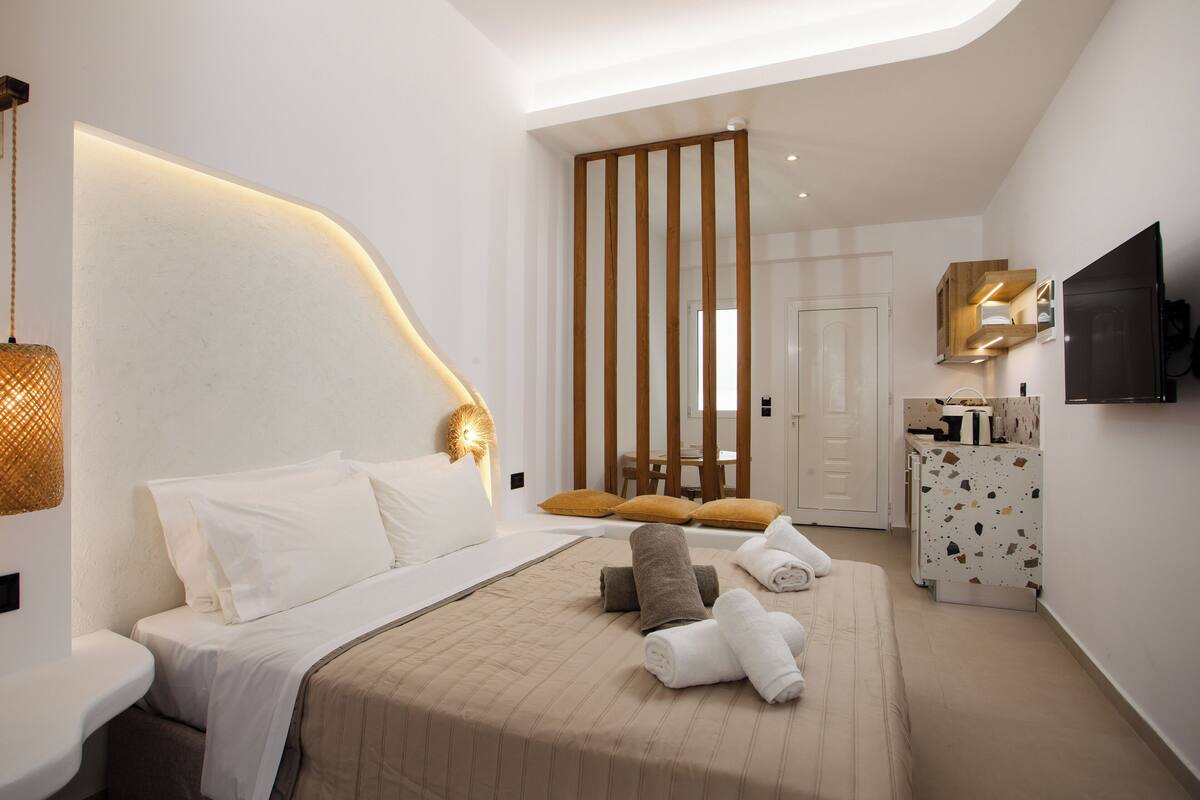 A modern hotel room with a large bed, a desk, a television, and a bathroom visible through a partition.