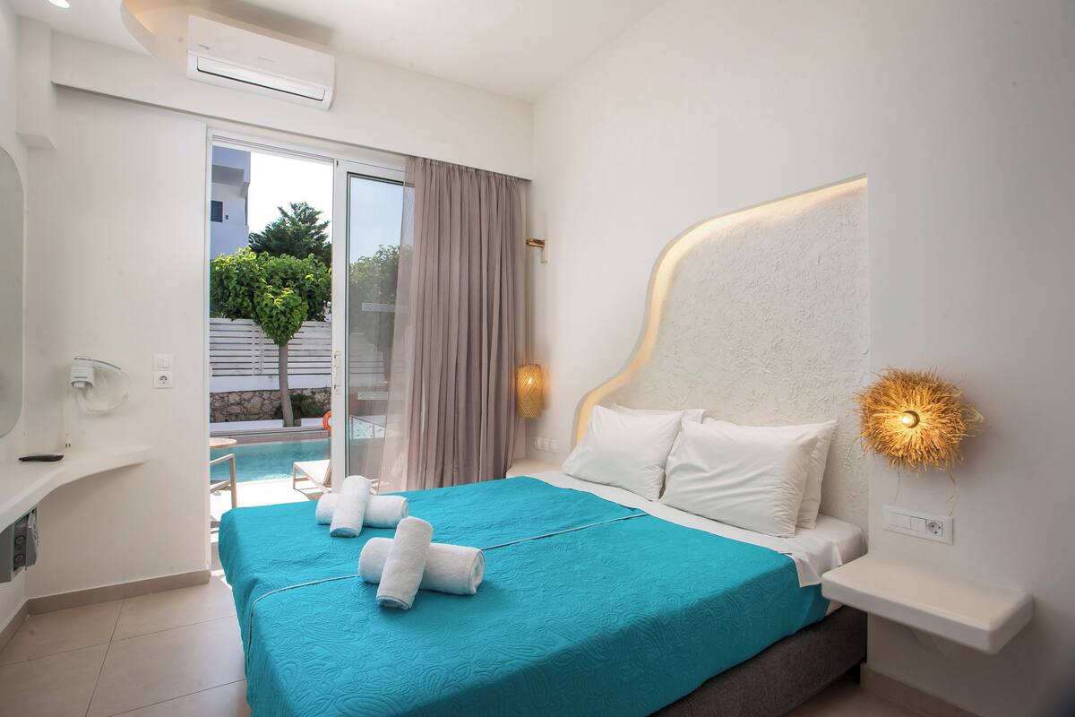 A modern hotel room with a bed, a bedside table, a nightstand, a built-in bench, a large window, and a view of a pool and greenery.
