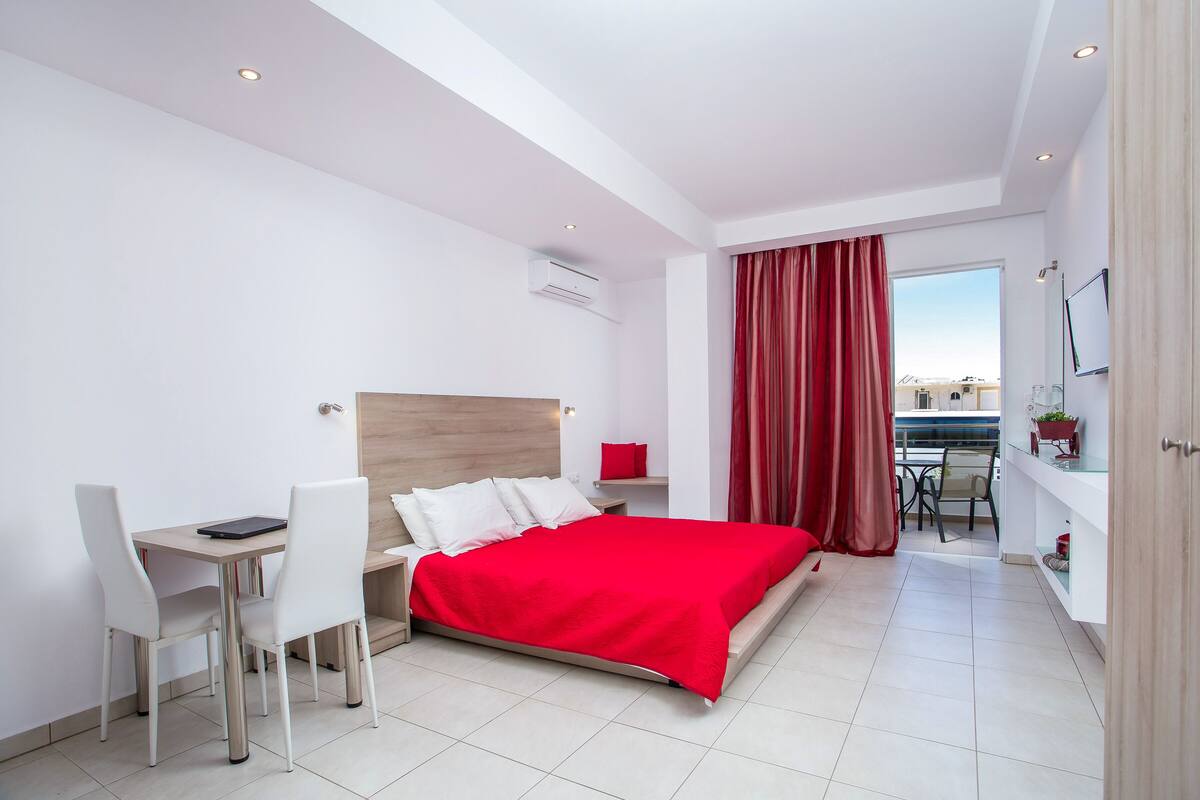 A modern hotel room with a bed, desk, and chair, a small dining area, and a view of the city.