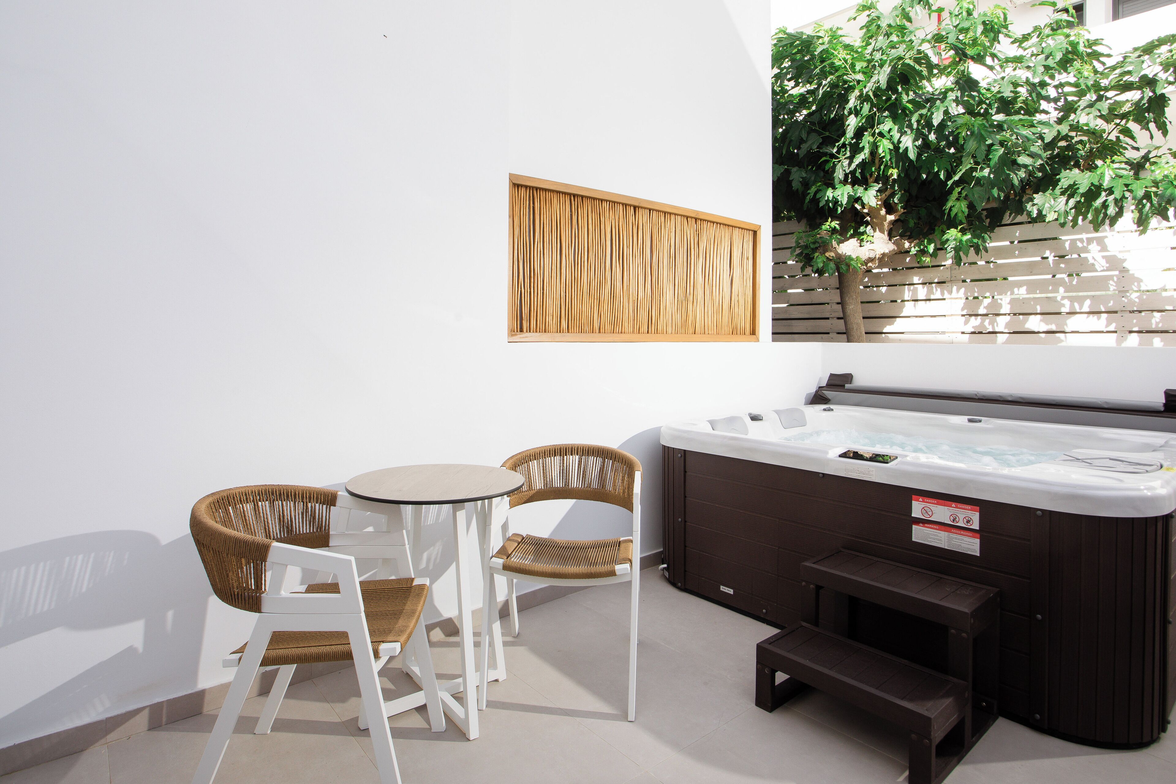 A hot tub area with two wicker chairs and a small table, a wooden slatted wall, and a large tree outside.