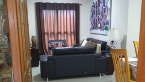 Una sala de estar con un sofá oscuro, un aparador de madera, una mesa de comedor con sillas y un cuadro abstracto grande en la pared.