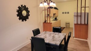 A dining area with a marble table and black chairs, a chandelier, a sunburst mirror, and wooden cabinets in the background.