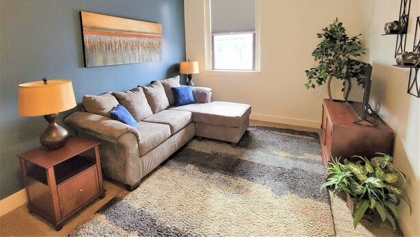 A living room with a sectional sofa, two side tables, a lamp, a painting, a window, and a potted plant.