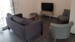 A modern living room with a grey sofa, a small round table, a television on a stand, and a grey armchair. - Beautiful 4-6 person gîte in the center of Vallon Pont d'Arc (Vallon-Pont-d'Arc)
