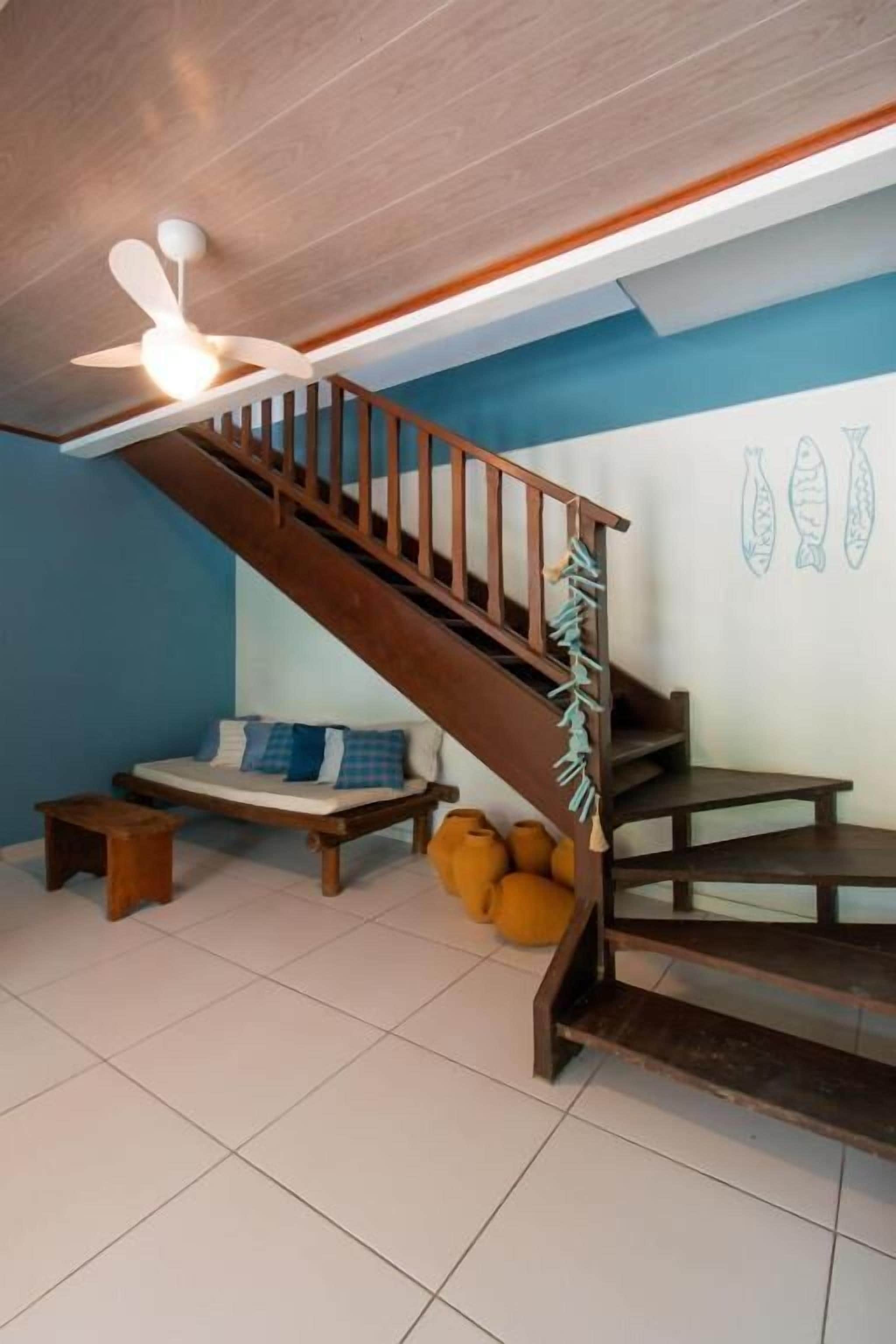 Un escalier en bois, un ventilateur de plafond, un canapé avec des coussins et un mur orné d’illustrations de poissons décoratifs.