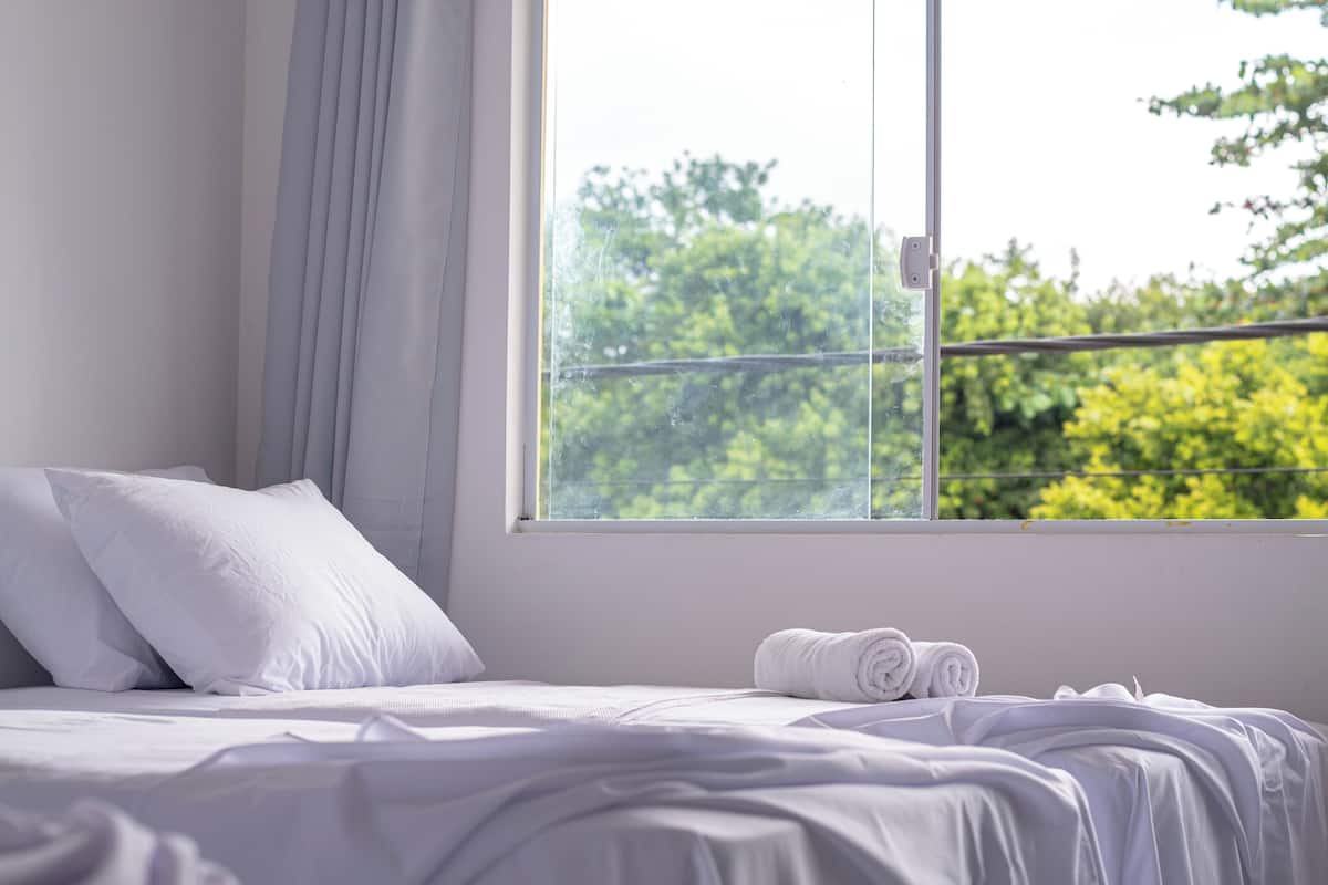 A neatly made bed with white linens and pillows, a window with a view of greenery, and a curtain partially drawn.