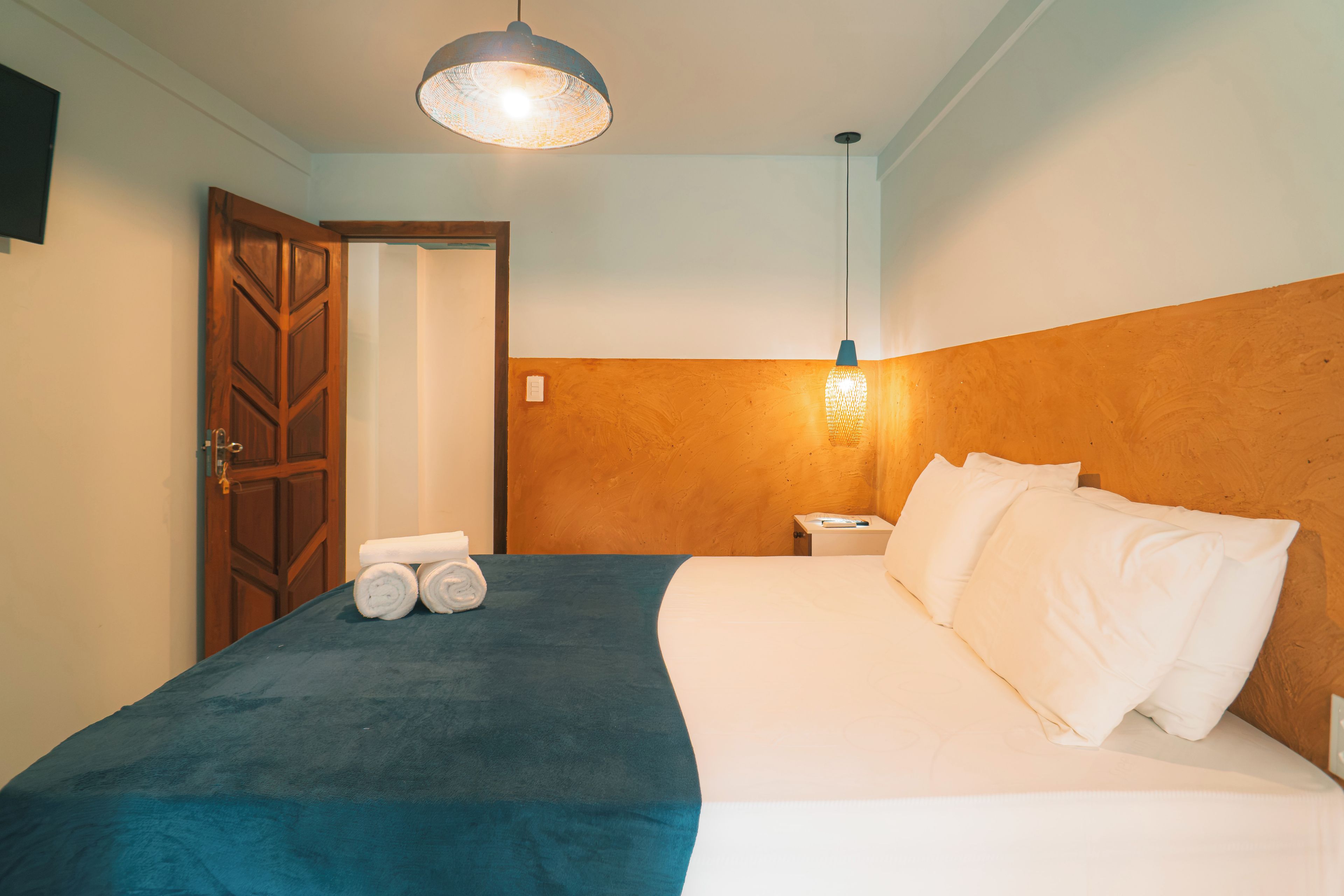 Quarto de hotel com cama, mesa de cabeceira, luminária na parede e uma porta.