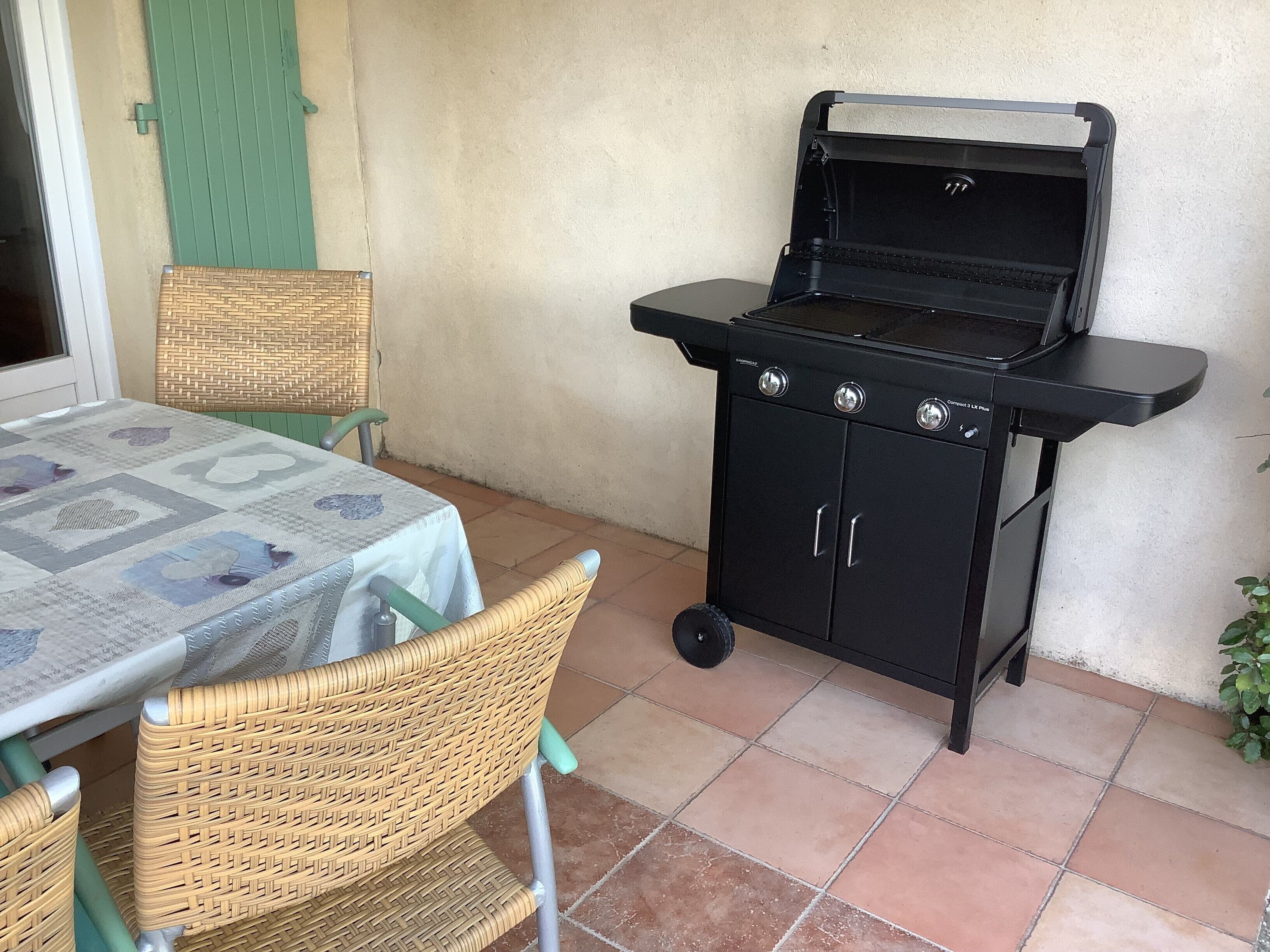 A patio with a table covered by a tablecloth, a wicker chair, a black barbecue grill, and a green door.