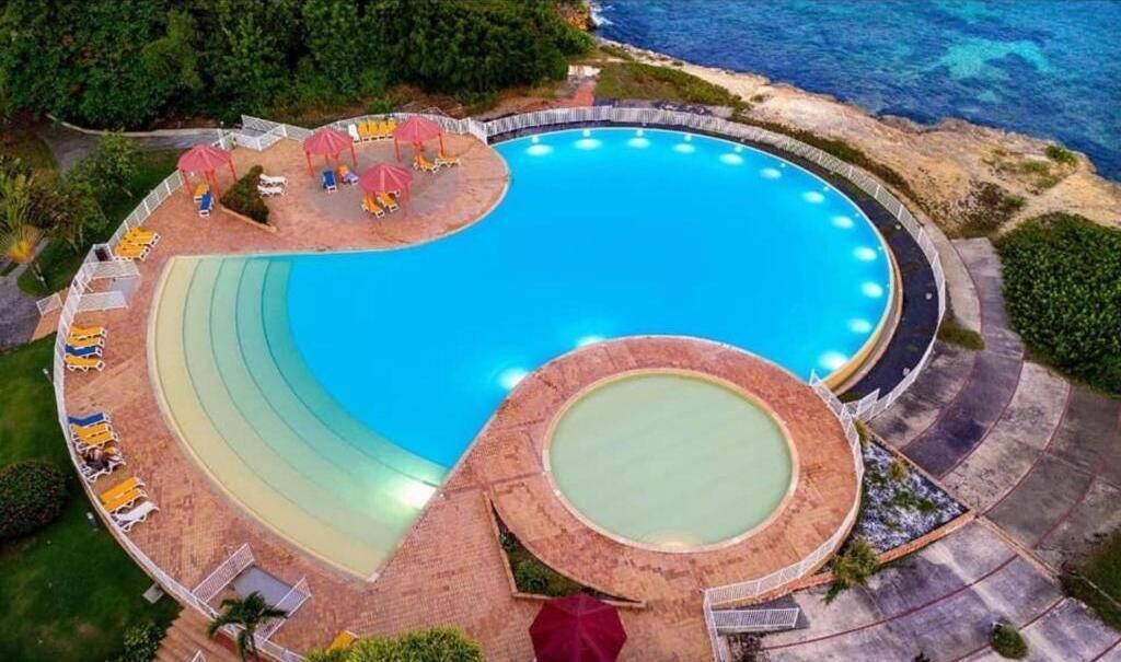 An aerial view of a resort with a large circular pool surrounded by lounge chairs and umbrellas, with a beach and ocean in the background.