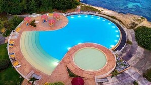 Ett flygbildsfoto av ett semesterområde med en stor, rund pool omgiven av solstolar och parasoller, med en strand och havet i bakgrunden.